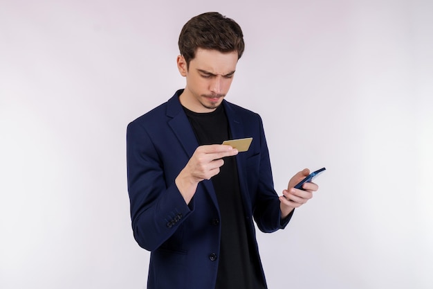 Portrait of unhappy young businessman standing using mobile cell phone and holding credit bank card isolated on white color background studio