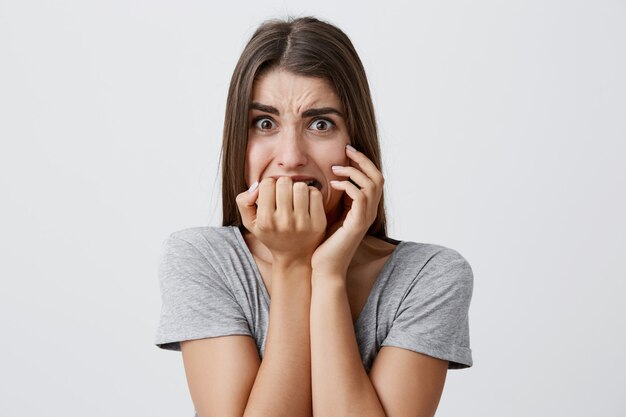 Portrait of unhappy young good-looking caucasian woman with long dark hair in casual gray t-shirt gnawing fingers,  with scared expression,