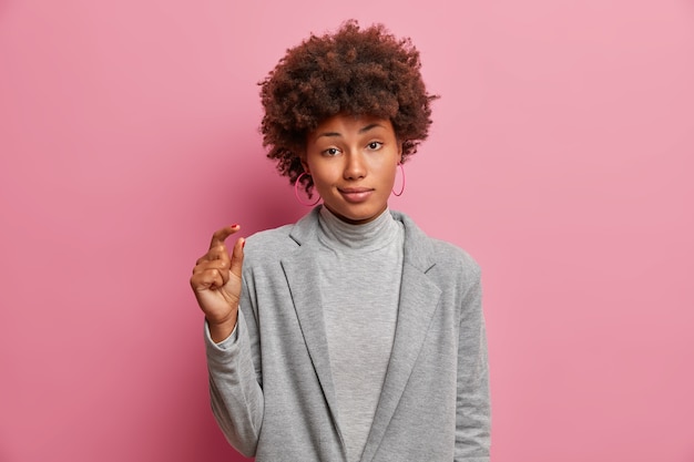 Free photo portrait of unimpressed curly woman raises hand and shapes tiny thing, shows very small object, dressed in grey outfit, tells about her poor salary