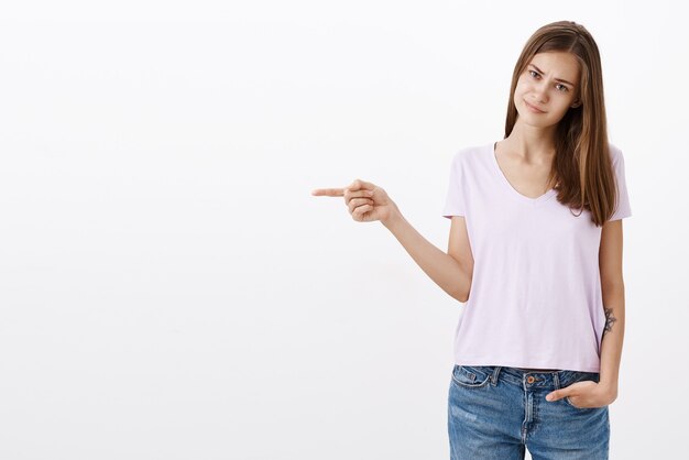 Portrait of unimpressed displeased cute snobbish girl with brown hair in t-shirt frowning and smirking from dislike pointing left standing dissatisfied and uninterested