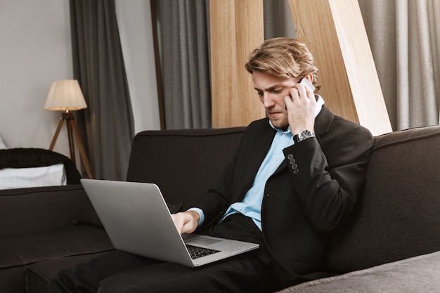 Portrait of unsatisfied young company director with trendy haircut and beard working with laptop computer, talking on phone with sales manager.