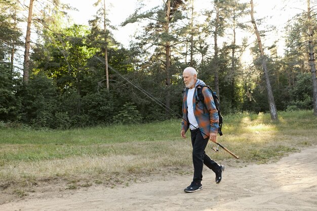 Portrait of unshaven bald European male pensioner with backpack, carrying fishery rod or spinning reel, going to catch fish on river bank. Recreation fishing, active healthy lifestyle and leisure