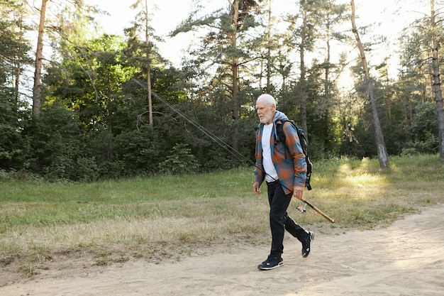 Портрет небритого лысого европейского пенсионера с рюкзаком, несущего удочку или спиннинговую катушку, собирающегося ловить рыбу на берегу реки. Рекреационная рыбалка, активный здоровый образ жизни и отдых