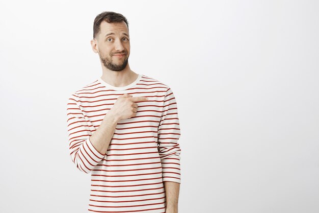 Portrait of unsure hesitating attractive man with beard, pointing right with index finger with displeased unimpressed expression