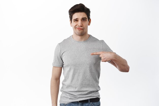 Portrait of unsure and skeptical adult man points at himself smirk and frown as look doubtful express disbelief standing over white background