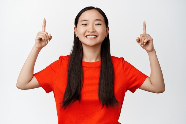 Portrait of upbeat smiling girl showing way, asian teen pointing fingers up and looking happy, announcing big sale, standing on white.