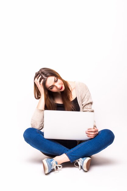 Portrait of an upset girl dressed in tank-top holding laptop while sitting on the floor over white