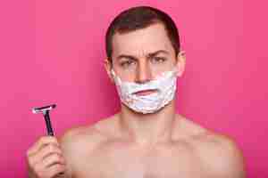 Free photo portrait of upset handsome athletic young man poses over bright pink wall in studio, looks dissatisfied on account of quality of razor and process of shaving. care and masculinity concept.