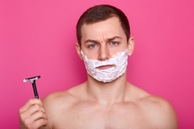 Portrait of upset handsome athletic young man poses over bright pink wall in studio, looks dissatisfied on account of quality of razor and process of shaving. Care and masculinity concept.