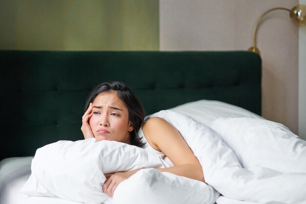 Portrait of upset korean woman asian girl lying in bed feels sad covers herself with duvet looks out