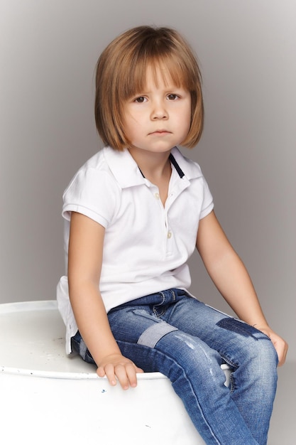 Portrait of an upset little girl in denim overalls, sitting in a studio on a gray background.