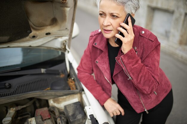 Portrait of upset middle aged European woman with gray short hair standing at her broken car with open hood because engine failure