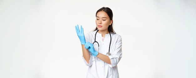 女性アジア人医師の肖像画は、健康状態に立っているクリニックで患者を調べるためにゴム手袋を着用します