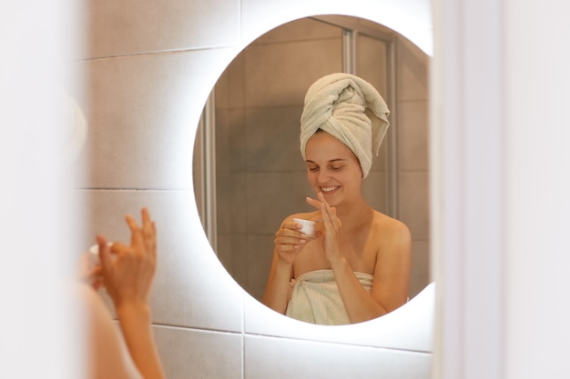 Portrait of woman being wrapped in white towel posing in bathroom in front of mirror open cream for applying on face, expressing happiness, skin care and cosmetology.