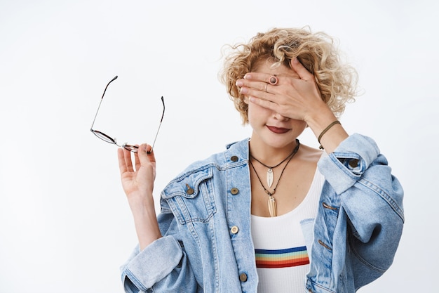 Portrait of woman cannot look at gross thing taking off glasses holding frames in hand as covering sight with palm and smiling waiting for time to open eyes over white wall