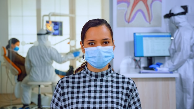 Foto gratuita ritratto di donna in studio dentistico che guarda la telecamera indossando la maschera facciale seduta su una sedia nella clinica della sala d'attesa mentre il medico lavora. concetto di nuova normale visita dal dentista nell'epidemia di coronavirus.