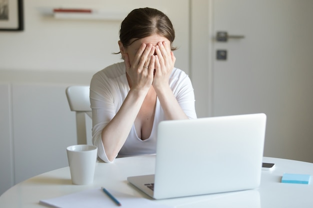 Portrait of woman at the desk with laptop, hands closing face