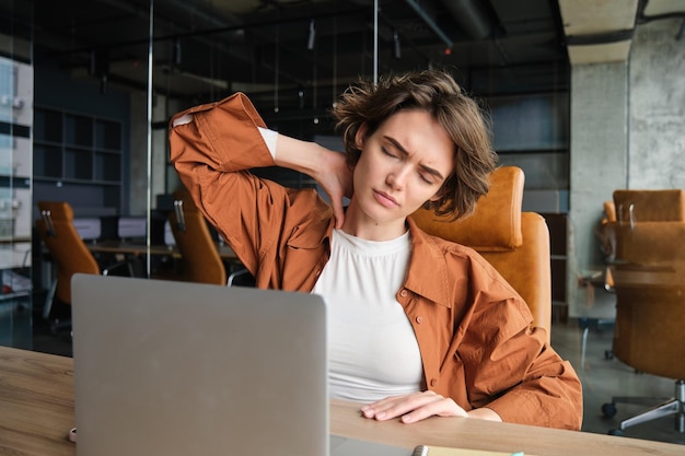 Foto gratuita ritratto di donna che sente dolore al collo e siede in ufficio con il portatile e ha tensione nei muscoli dopo il lavoro