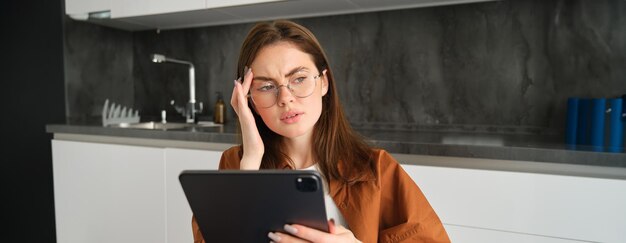 Portrait of woman freelancer wearing glasses holding digital tablet looking tired has migraine or