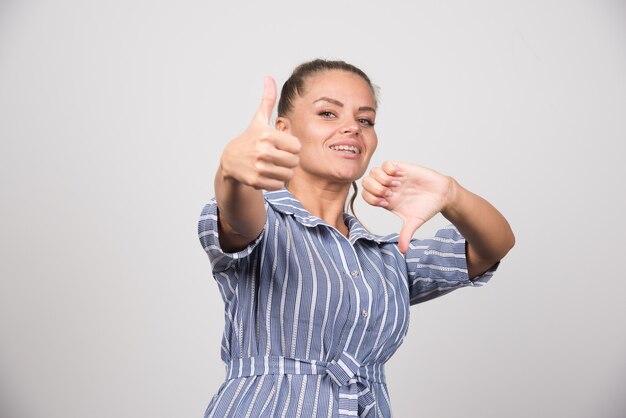 Portrait of woman giving thumbs up and down on gray wall.