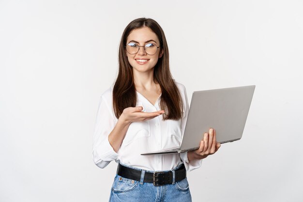 Portrait of woman in glasses holding laptop pointing at screen showing her work on computer standing...