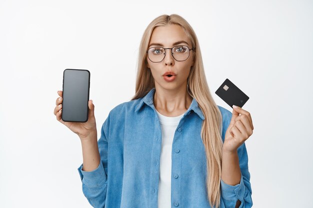 Portrait of woman in glasses reacting surprised showing mobile phone screen with credit card staring impressed at camera white background