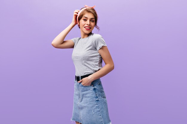 Portrait of woman in grey T-shirt posing on purple background. Wonderful young girl in denim short skirt with black wide belt smiling.