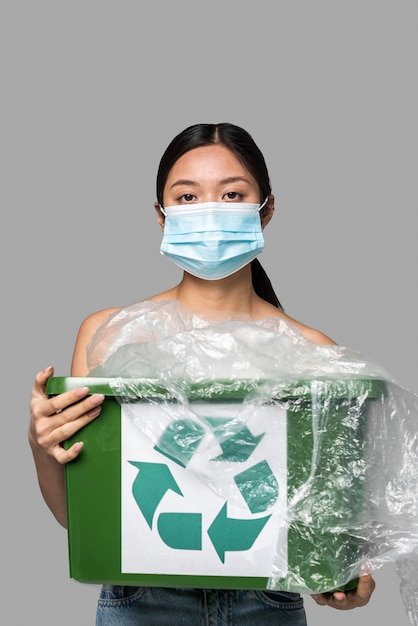 Free photo portrait of woman holding a recycle bin while wearing a medical mask