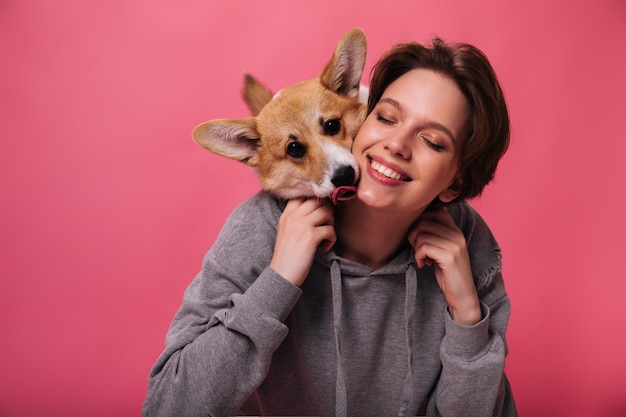 Portrait of woman in hoodie hugging her dog on pink background