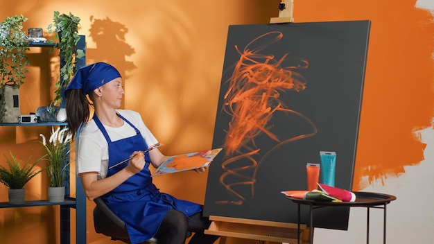 Free photo portrait of woman painting artwork on canvas with brush, using artistic tools and watercolor aquarelle on mixing tray. creating colorful masterpiece with orange wet dye and paintbrush. tripod shot.