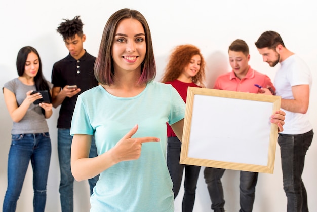 Portrait of woman pointing towards blank white frame while her friends busy in cellphone