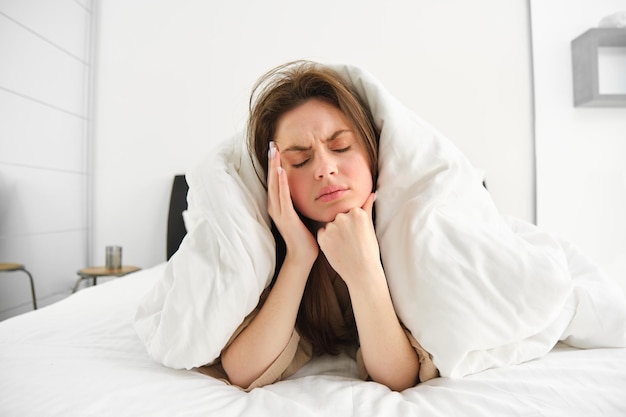 Portrait of woman rolled in blankets in bed frowning and feeling unwell has hangover headache in