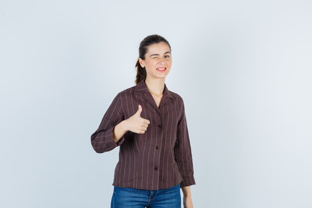 Portrait of woman showing thumb up while blinking in shirt, jeans and looking happy front view