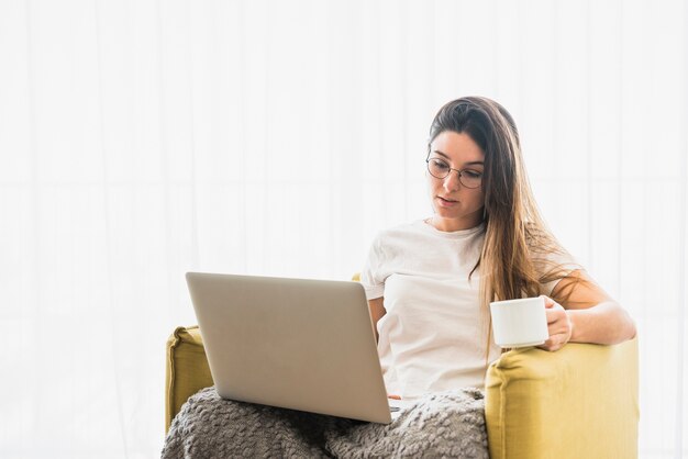 Portrait of woman sitting on armchair holding coffee cup using laptop