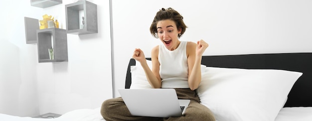 Free photo portrait of woman sitting on bed looking at laptop with excited amazed face celebrating making fist