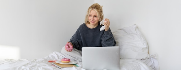 Portrait of woman sitting on a bed with laptop eating doughnut and crying from sad movie scene