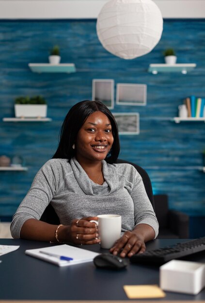 Portrait of woman smiling and sitting at desk with computer, working from home on business project. Young adult looking at camera and using monitor with keyboard to do remote work.