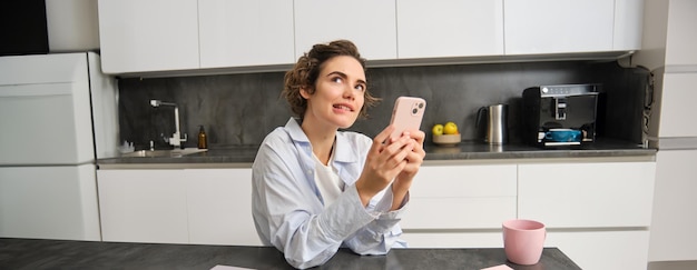 Free photo portrait of woman thinking while holding smartphone deciding what to order on mobile phone app