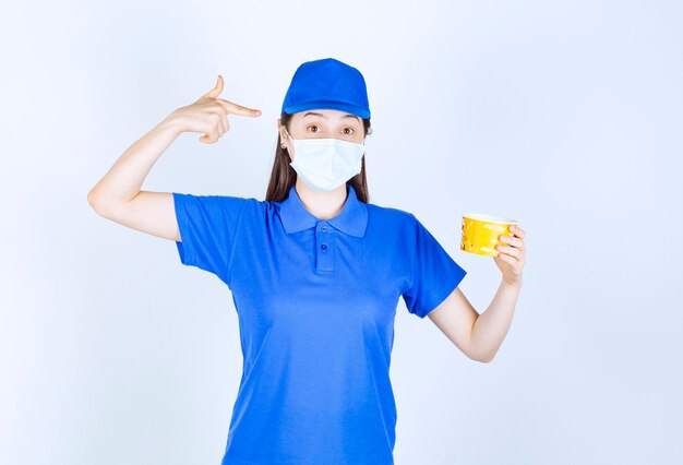 Portrait of woman in uniform and medical mask pointing at plastic cup