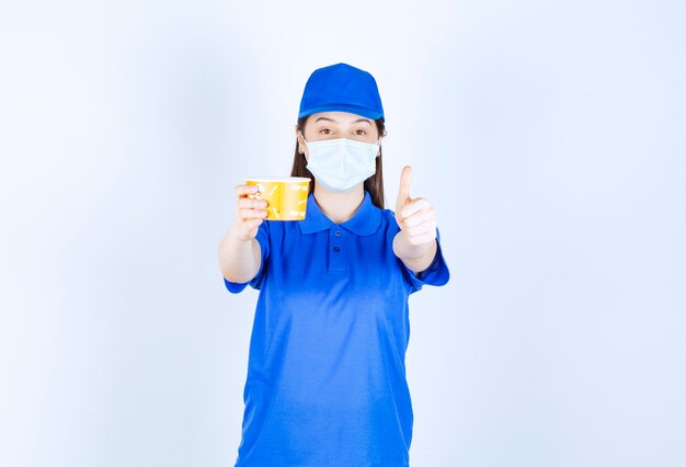 Portrait of woman in uniform and medical mask with plastic cup showing thumb up