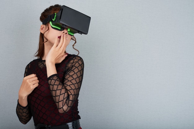 Portrait of woman in VR goggles touching her lips standing against grey