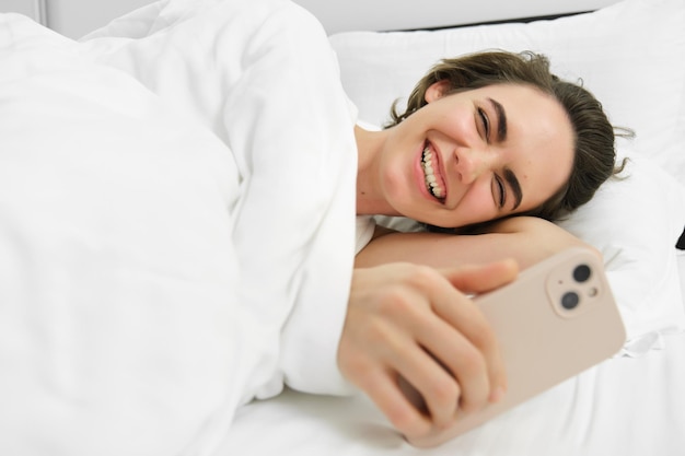 Foto gratuita ritratto di una donna che guarda video divertenti nel suo letto guardando lo smartphone e ridendo usando il cellulare