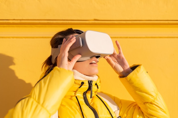 Free photo portrait of a woman wearing virtual reality glasses on the street against a yellow wall in bright sunlight in winter in warm clothes