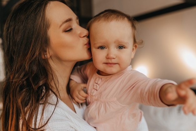 Portrait of woman in white home clothes and her blue-eyed baby. Lady lovingly kisses her daughter.