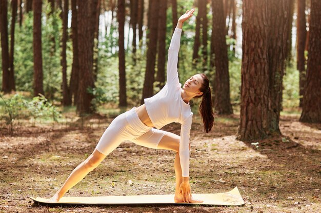 Portrait of woman in white stylish sport top and leggins standing on mat in yoga position in beautiful forest, stretching body, practicing yoga outdoors.