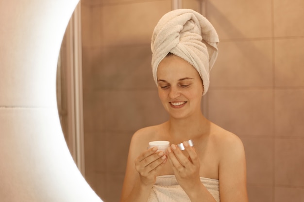 Портрет женщины с кремом в руках, отражение улыбающейся женщины с полотенцем на голове, делающей косметические процедуры дома в ванной, заботясь о ее коже.