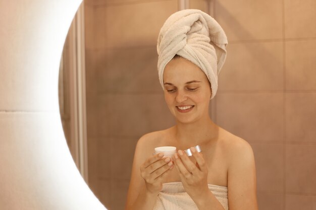 Portrait of woman with cream in hands, reflection of smiling female with towel on her head doing beauty procedures at home in bathroom, taking care of her skin.