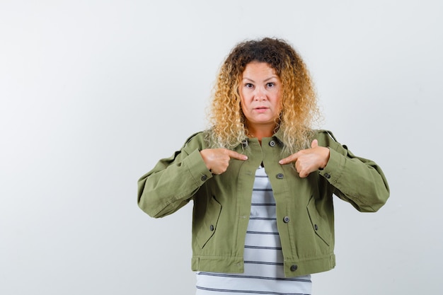 Portrait of woman with curly blonde hair pointing at herself in green jacket and looking perplexed front view