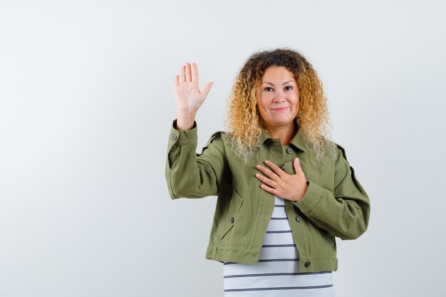 Portrait of woman with curly blonde hair showing palm, keeping hand on chest in green jacket and looking grateful front view
