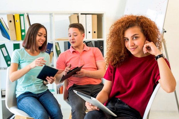 Portrait of a woman with curly hairs sitting on chair holding digital tablet with her friends at background